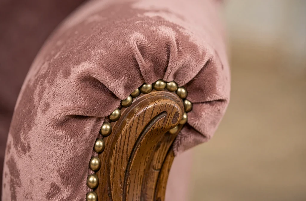 Détail de l'accoudoir d'un fauteuil Voltaire retapissé en velours rose avec garniture de clous dorés